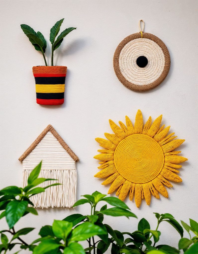 A collection of woven wall hangings featuring a bright yellow sun and a traditional-style potted plant, mounted on a neutral wall with greenery in the background.
